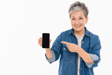 Excited mature gray-haired woman in casual outfit denim shirt pointing index finger on mobile cell phone with blank empty mock up screen isolated on white background studio portrait copy space