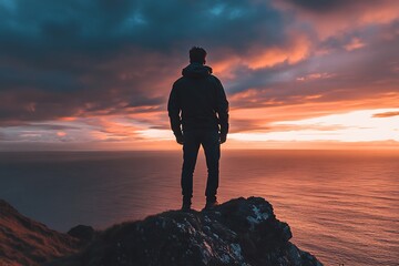 Silhouette of a man standing on cliff overlooking ocean and dramatic sunset sky