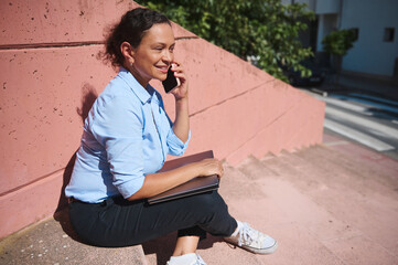 Smiling young woman talking on phone while sitting on outdoor steps in casual attire, holding a laptop, enjoying a break in sunny weather