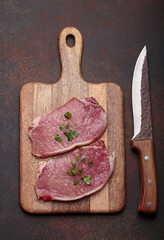 Two raw pork steaks are seasoned with spices and ready for cooking on a wooden chopping board. A sharp knife lies beside the meat, ready to be used