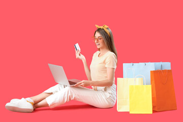 Wall Mural - Beautiful happy stylish young woman with shopping bags, credit card and laptop sitting on red background