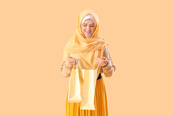 Young beautiful woman in hijab opening shopping bags on pale orange background