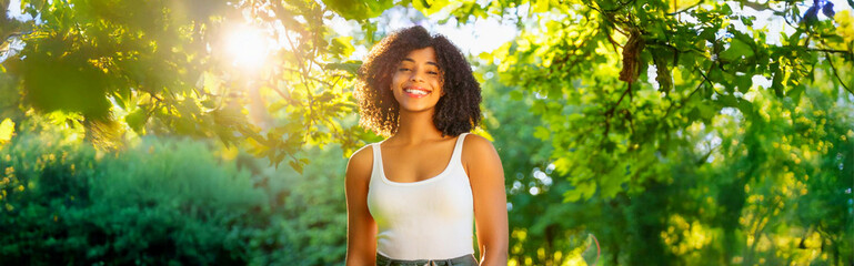 A portrait of a Black woman wearing a white tank top, smiling in the forest.