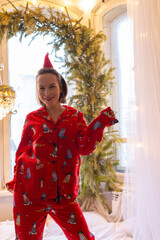 Happy girl in red pajamas dancing and jumping on bed alone at home in light room. Young woman ejoying Christmas time, vertical shot. Positive emotions on Xmas holiday