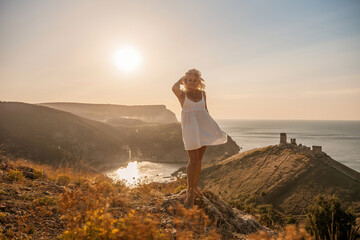 A blonde woman stands on a hill overlooking the ocean. She is wearing a white dress and she is enjoying the view.