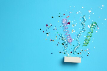 Canvas Print - Shiny confetti and party streamers flying out of little box on light blue background, flat lay. Space for text