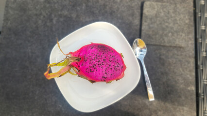 A halved dragon fruit lies on a white plate. The flesh is bright pink and studded with numerous small black seeds.