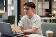 © bongkarn - A handsome, focused Asian man working remotely from a coffee shop, working on his laptop.