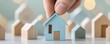 © ParinApril - A hand placing a blue model house among smaller wooden houses, symbolizing home selection and real estate concepts.