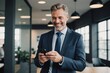 © ThomasLENNE - smiling happy confident mid aged male company ceo executive wearing suit holding cellphone standing in office using business mobile apps technology financial online solutions on cell phone