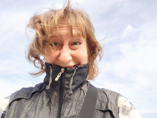 A middle-aged female tourist blogger takes a selfie on a travel. A woman wearing a gray vest and plaid shirt smiles for a selfie on a sunny day