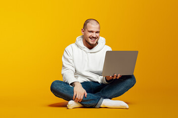 Portrait of a happy young bearded man in white hoodie holding laptop computer while sitting on a floor isolated over yellow background.