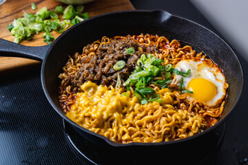 cooking savory ramen noodles topped with beef slice, egg and cheese in the cast iron skillet, with a spring onion as a garnish, kitchen setting