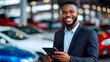 © Tanya - Smiling African salesman holding a digital tablet in an auto showroom, surrounded by various cars, providing excellent customer service. Copy space.
