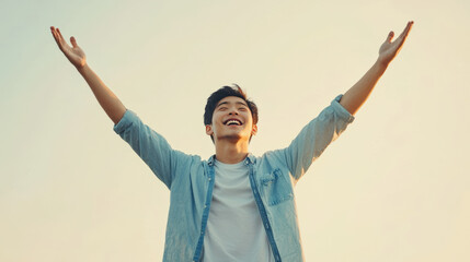 A joyful young man in light blue shirt raises his arms in celebration against clear sky, expressing happiness and freedom