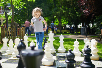 Adorable preschooler girl playing chess with big chess pieces outdoors on a sunny summer day.