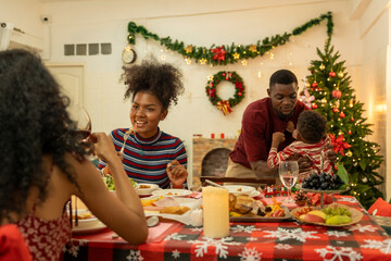 A joyful family gathers around the dinner table celebrating Christmas. The father, dressed in a Santa hat, gives a gift to the child, while everyone smiles with warmth and festive decorations.