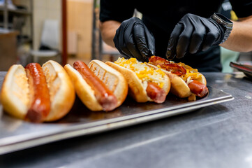 Wall Mural - Close-up hands preparing hot dogs with various toppings on stainless steel surface, showcasing fast-food concept. Perfect for illustrating culinary themes, street food, and casual dining experiences.