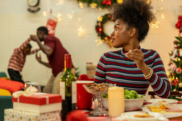 A joyful family gathers around the dinner table celebrating Christmas. The father, dressed in a Santa hat, gives a gift to the child, while everyone smiles with warmth and festive decorations.