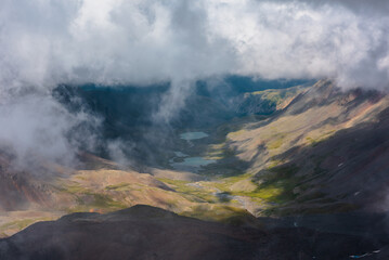 Aerial top view to green rocky hilly valley with two turquoise lakes under lush low clouds. Most beautiful mountain lake under thick low cloud. Sunlight and shadows in mountains in changeable weather.