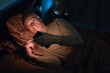 © Andrii Lysenko - Pensioner old man uses mobile phone lying in his bed at night suffering from insomnia and mobile addiction