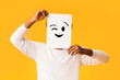 © Prostock-studio - Black guy holding paper with positive emoji over orange background, hiding his real emotions
