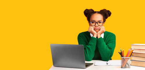 Bored Teenager. Portrait of sad black girl using laptop sitting at table isolated on yellow wall background, copy space