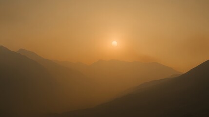 Poster - A golden sun sets behind rugged mountain silhouettes, casting a warm glow in the hazy atmosphere of late evening. The landscape is filled with soft shadows and tranquil colors as dusk approaches.