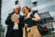 © qunica.com - Two business colleagues discussing and reviewing documents outside a modern office building. They're engaged in conversation and collaboration.