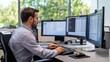 © Elmira - A professional man types intently at his desk, engaged with three computer monitors displaying data in a bright, contemporary office with large windows