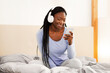 © Prostock-studio - Weekend Morning. Cheerful Afro Girl In Headphones Using Smartphone Listening To Music Sitting In Bed In Cozy Bedroom