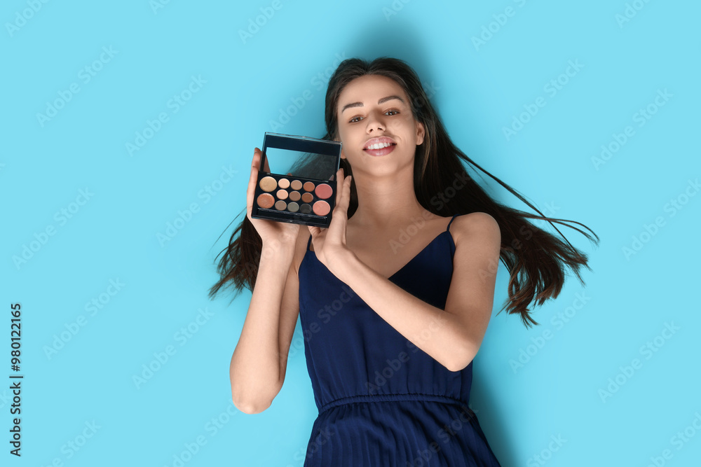 Happy smiling young woman with eyeshadows palette lying on blue background
