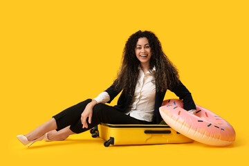 Poster - Happy female office worker with inflatable ring and suitcase sitting on yellow background. Summer vacation concept