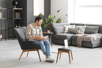 Canvas Print - Young male teacher with laptop, book and notebook giving online lesson at home