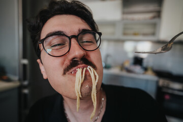 Close-up of a man wearing glasses and mustache, playfully eating spaghetti. The scene captures humor and enjoyment in a casual kitchen setting.