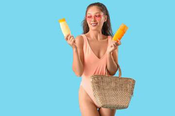 Wall Mural - Young Asian woman in swimsuit with bottles of sunscreen cream and beach bag on blue background