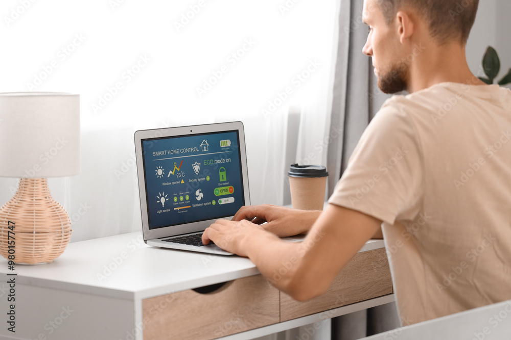 Young man using laptop with smart home application at table in room