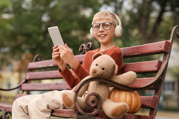 Wall Mural - Cute boy with headphones and pumpkin using tablet computer on bench in park