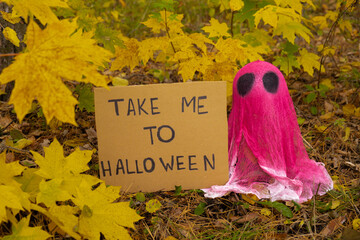 Creative handmade Halloween holiday decoration. pink ghost with cardboard message TAKE ME TO HALLOWEEN in autumn leaves forest. Zero waste homemade decor. Mystic place supernatural 