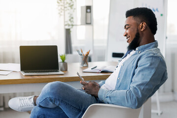 Poster - Idleness in office. Carefree black employee using smartphone during working hours browsing internet or typing message, side view
