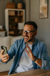 © La Famiglia - Excited Man Using Smartphone for mobile shopping while Sitting in Living Room. Happy Man buying things, shopping online, using a mobile app