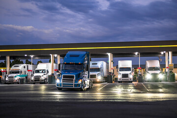 Commercial big rig semi trucks with semi trailers fill up their fuel tanks at truck stop gas station at night time to continue driving on their routes in the morning