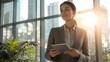 © CStock - A confident young business woman dressed in a modern, professional suit, standing by a large window in a bright office, holding a tablet,