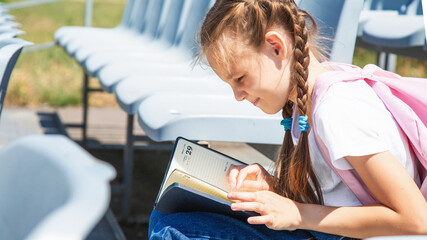 Elementary school girl or school pupil is studying, preparing to lesson, reading book or textbook outdoors at campus with backpack