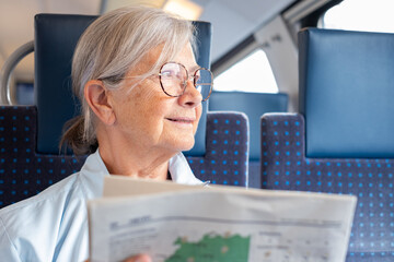 Senior woman enjoys the train ride sitting in passenger seat by the window with a newspaper in hand, smiling elderly woman savors the time spent on the journey by reading