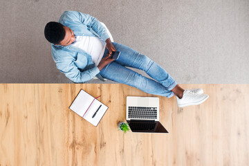 Poster - Idleness at work. Top view of carefree afro employee using smartphone and keeping his legs on table during working day in office, copy space