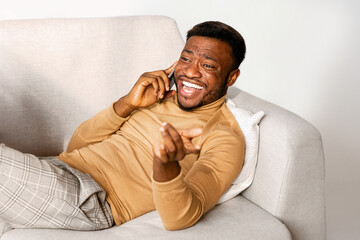 Canvas Print - Joyful African American Guy Talking On Cellphone And Laughing Holding Laptop Computer Lying On Sofa Indoor. Selective Focus