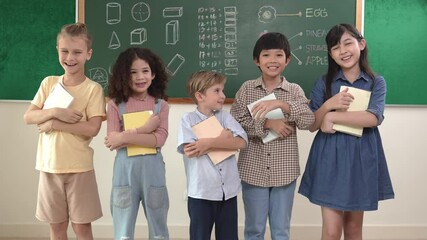 Wall Mural - Multicultural elementary student in casual cloth looking at camera while standing at classroom. Group of cute diverse children hold notebook and smiling to camera at blackboard in classroom. Pedagogy.