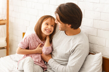 Wall Mural - Cute little girl playing with her young father at home, sitting on bed, dad tickling daughter, copy space