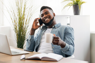 Poster - Coffee break. Cheerful african american employee talking on phone and holding cup with hot drink while resting after successful work in office.